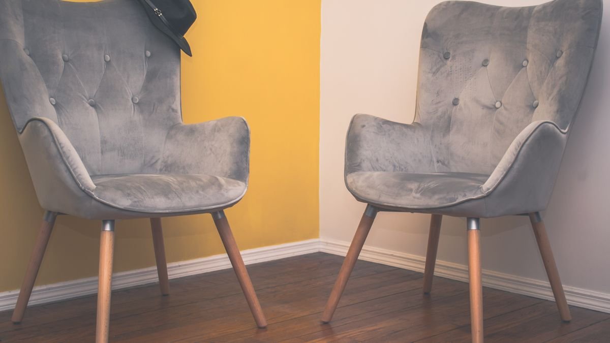 Two contemporary gray armchairs in a minimalist interior with a yellow accent wall and wooden flooring.