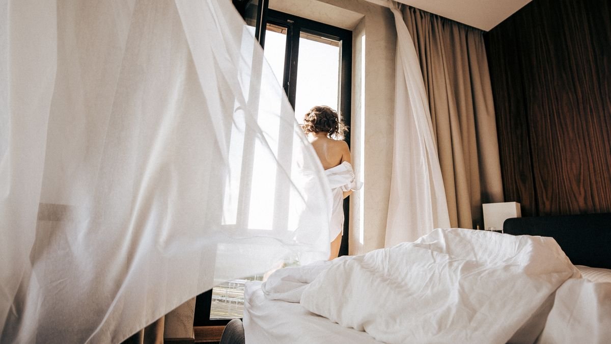 A person stands by an open window in a bedroom with flowing curtains and soft morning light.