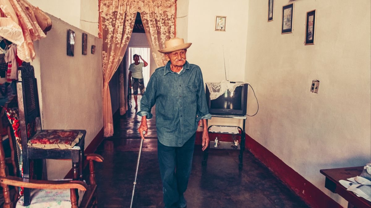 Senior man with a cane walks indoors, surrounded by vintage furniture and decor.
