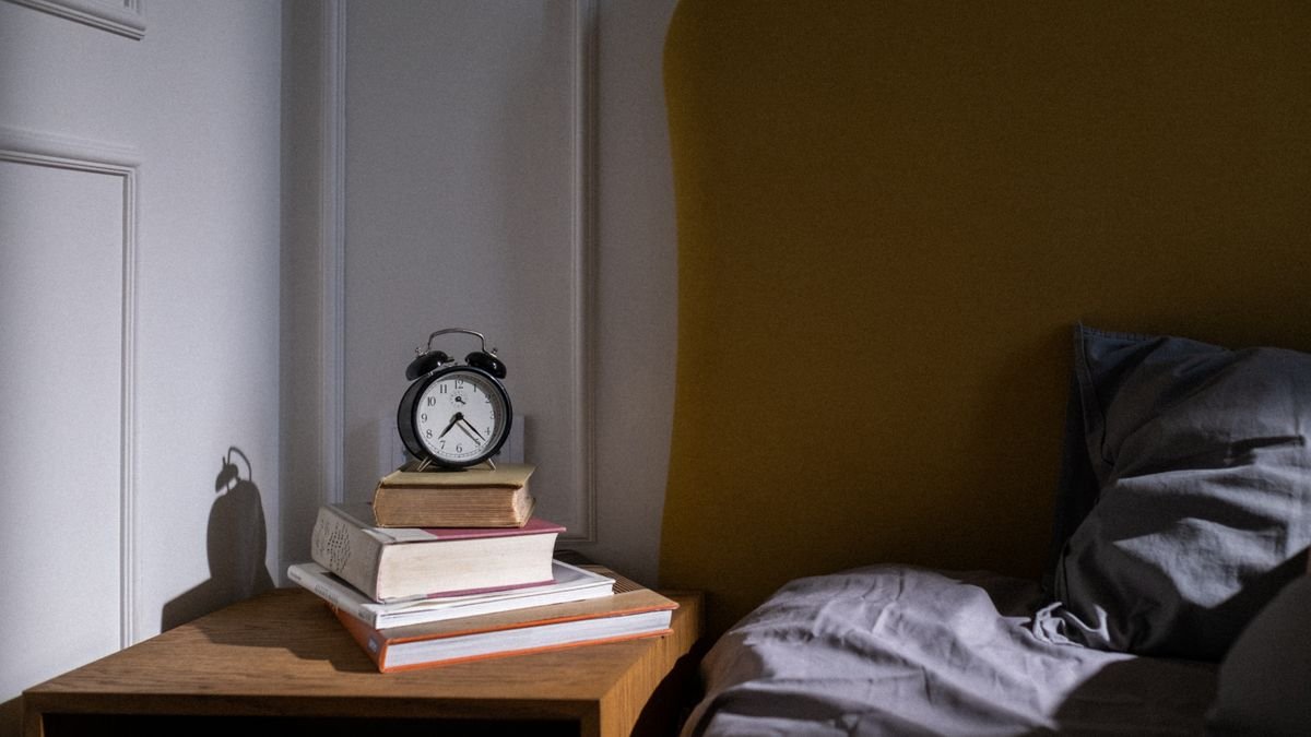 A cozy bedroom setting with books and an alarm clock on a nightstand next to a bed.