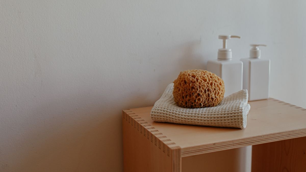 Close-up of a bathroom shelf featuring a natural sponge, towels, and pump bottles.